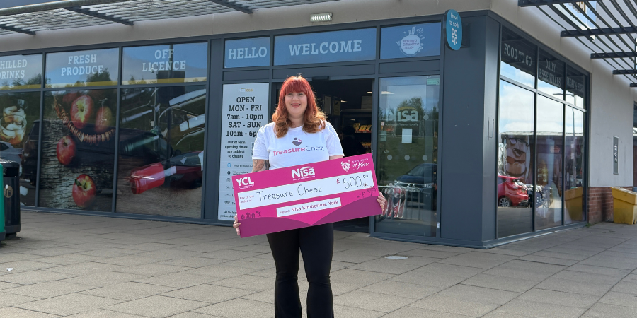 A woman standing in front of a Nisa Local storefront, holding a large ceremonial charity cheque for £500 made out to 